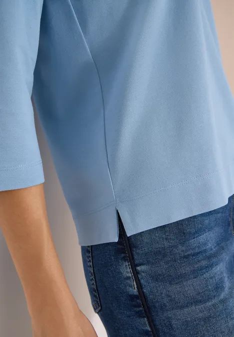 Polo à manches 3/4 avec boutons décoratifs easy blue Person trägt hellblaues Shirt mit 3/4-Ärmeln und seitlichem Saumschlitz, kombiniert mit dunkler Jeans.