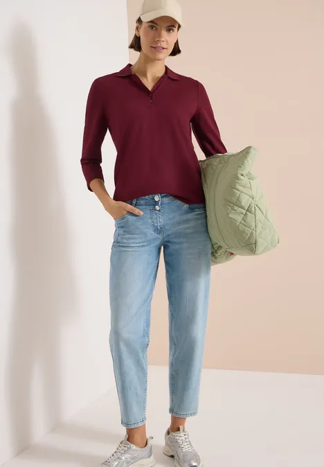 3/4-Arm Poloshirt mit Knopfdetail red wine Frau trägt eine grüne Stepp-Tasche, bordeauxrote Bluse und helle Jeans vor hellem Hintergrund.