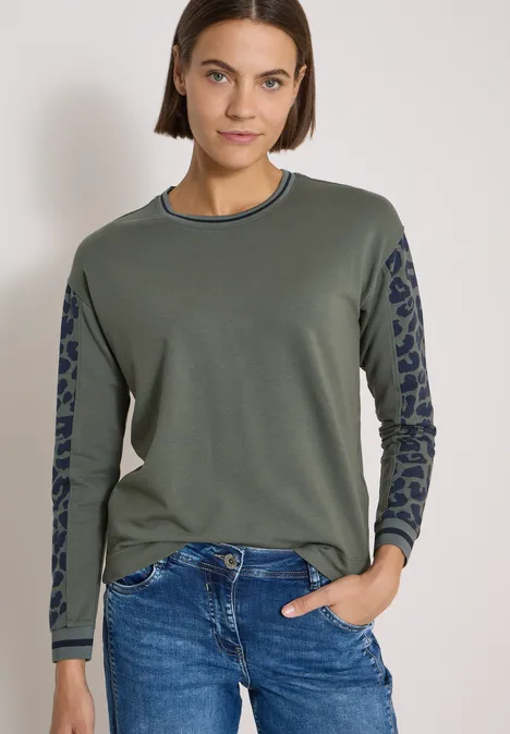 Frau mit braunen Haaren trägt olivgrünen Pullover mit gemusterter Passe an den Ärmeln und blaue Jeans.