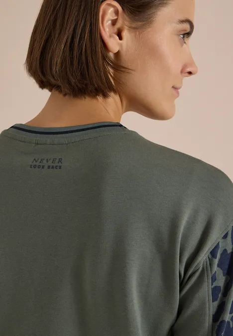 Rückenansicht einer Frau mit kurzem Bob, grüner Sweatshirt mit Musterärmel und Schriftzug „Never Look Back“.