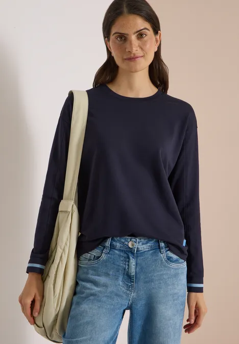 T-shirt à manches longues, col rond et rayures urban dark blue Frau in dunkelblauem Pullover und Jeans mit Beuteltasche vor neutralem Hintergrund.