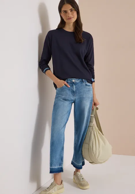 T-shirt à manches longues, col rond et rayures urban dark blue Frau mit dunkelblauem Shirt, lockeren Jeans und weißen Sneakern lehnt lächelnd an Wand und trägt beige Tasche.