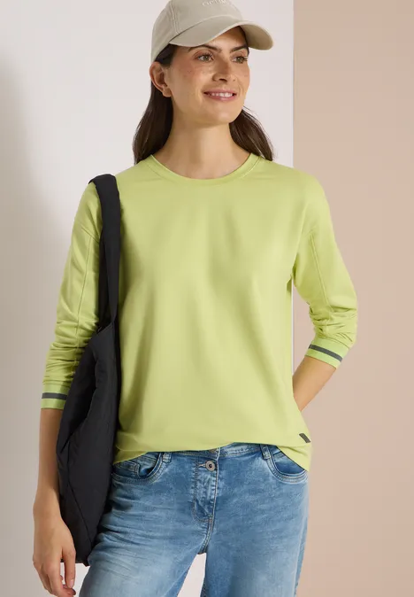 T-shirt à manches longues, col rond et rayures citrin green Fröhliche Frau mit hellgrünem Langarmshirt, Jeans, Baseballmütze und schwarzer Tasche vor neutralem Hintergrund.