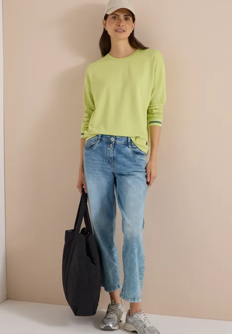 T-shirt à manches longues, col rond et rayures citrin green Frau trägt grünes Shirt, Jeans, Cap, Sneakers, hält schwarzen Shopper, lächelt vor beiger Wand.