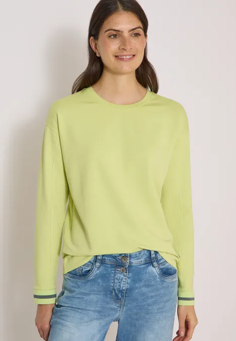 T-shirt à manches longues, col rond et rayures citrin green Junge Frau mit limettengrünem Pullover und Jeans, lächelnd vor hellem Hintergrund.