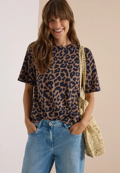 Frau in Leoparden-Shirt und Jeans lächelt mit handgewebter Tasche vor neutralem Hintergrund.