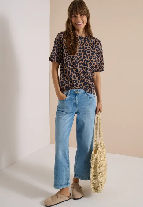 Frau trägt Leo-Shirt, weite Jeans und Basttasche, lächelt