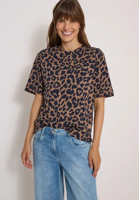Lächelnde Frau in Leo-T-Shirt und Jeans vor heller Wand.