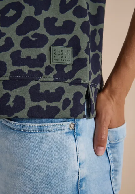 Grau-grünes T-Shirt mit Camouflage-Muster, seitlichem Schlitz und Label-Patch über hellblauen Jeans.