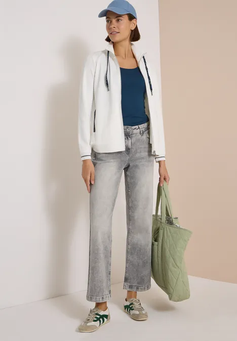 Frau mit blauer Mütze, weißer Kapuzenjacke, grauen Jeans und grüner Tasche vor neutraler Wand.