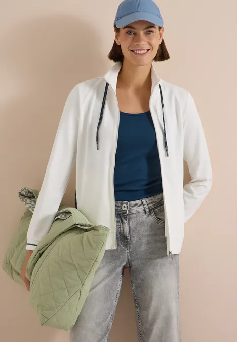 Frau in weißer Jacke, blauem Top, Jeans und Cap hält grüne gesteppte Jacke und lächelt.