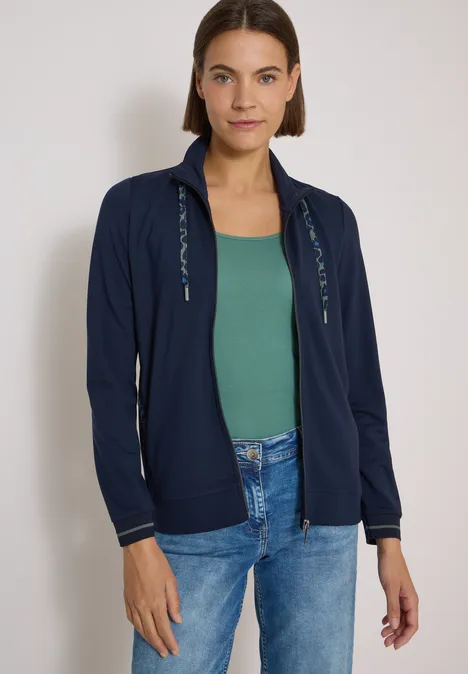 Eine Frau mit kurzen braunen Haaren, dunkelblauer Kapuzenjacke, grünem Top und Jeans steht vor hellem Hintergrund.