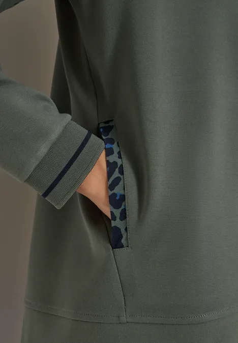 Hand in Seitentasche eines olivegrünen Sweatshirts mit camouflagefarbenem Besatz und dunkelblauem Saum.
