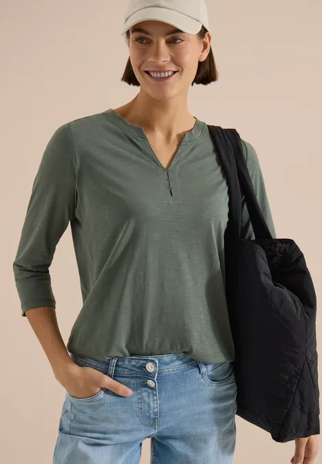3/4-Arm Shirt mit Split Neck und Elastiksaum dried olive green Eine lächelnde Frau in grüner 3/4-Arm-Bluse, Jeans und weißem Cap mit schwarzer Umhängetasche.