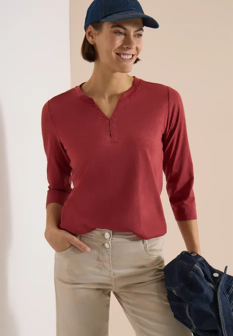 3/4-Arm Shirt mit Split Neck und Elastiksaum russet red Frau trägt rotes Langarmshirt mit Knopfleiste und helle Hose, hält lässig eine Jacke in der Hand.