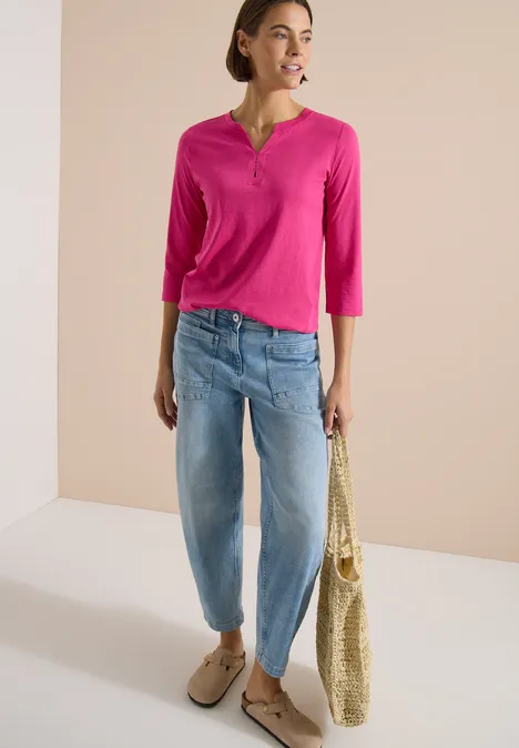 3/4-Arm Shirt mit Split Neck und Elastiksaum pink flash Frau trägt pinkes Henley-Shirt, helle Boyfriend-Jeans, Basttasche und beige Loafers vor hellem Hintergrund.