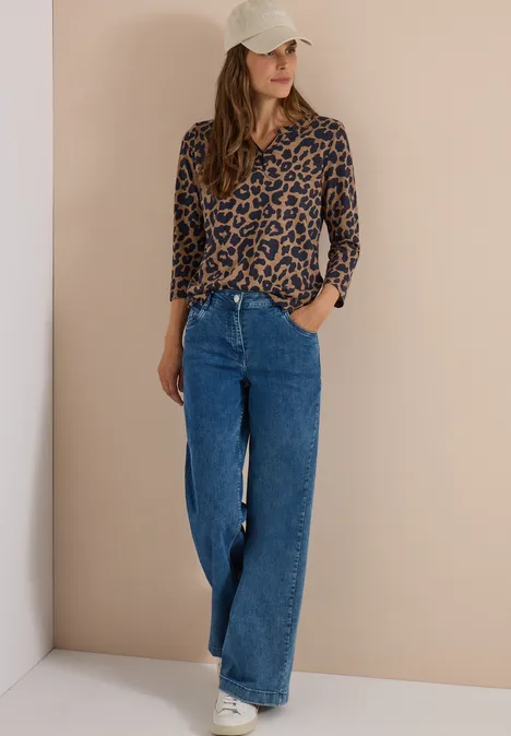 Frau mit Leopardenmuster-Bluse, weiten Jeans, Baseballcap und Sneakern steht vor beiger Wand.