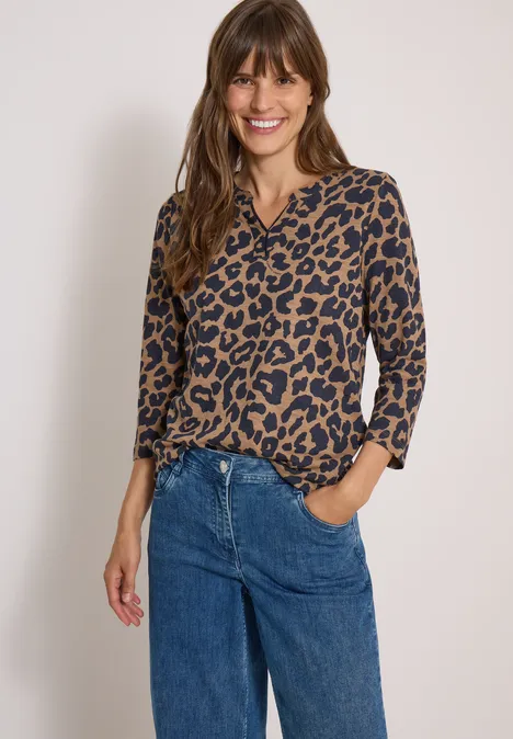 Frau trägt eine Leo-Bluse und blaue Jeans, lächelt entspannt mit einer Hand in der Hosentasche.