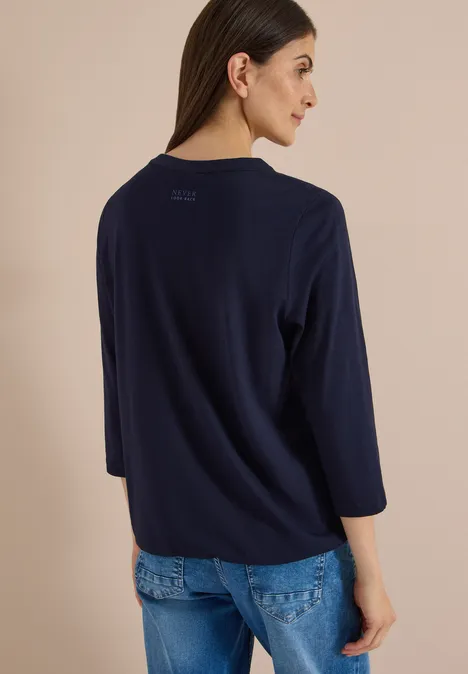 Frau in dunkelblauem Pullover mit 3/4-Ärmeln und Jeans, Rückenansicht vor hellem Hintergrund.
