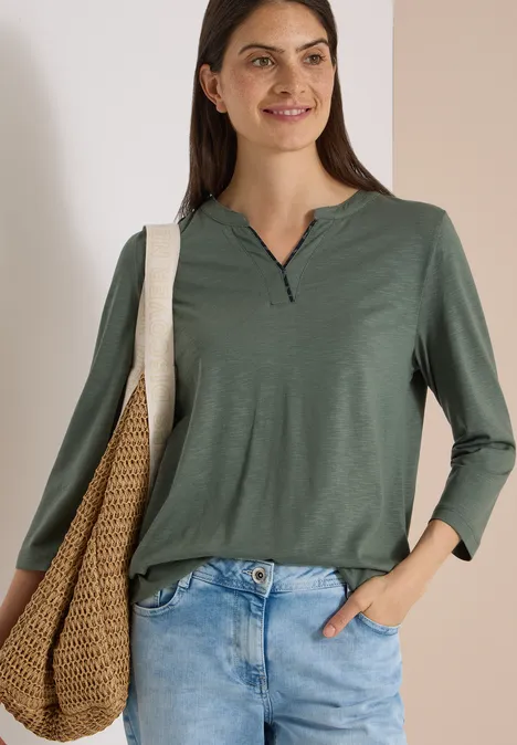 3/4-Arm Shirt mit Split Neck dried olive green Frau in grünem Longsleeve, trägt Jeans und Korbtasche, lächelt vor hellem Hintergrund.
