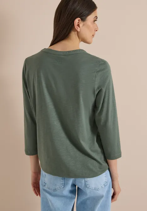 3/4-Arm Shirt mit Split Neck dried olive green Frau in olivgrünem 3/4-Arm-Shirt und hellblauen Jeans, Rückenansicht vor beigem Hintergrund.