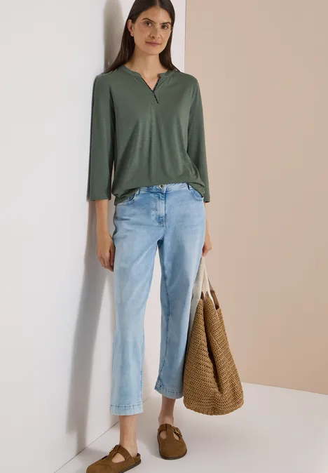 3/4-Arm Shirt mit Split Neck dried olive green Frau trägt grünes Shirt, helle Jeans, braune Hausschuhe, hält große geflochtene Tasche vor neutraler Kulisse.