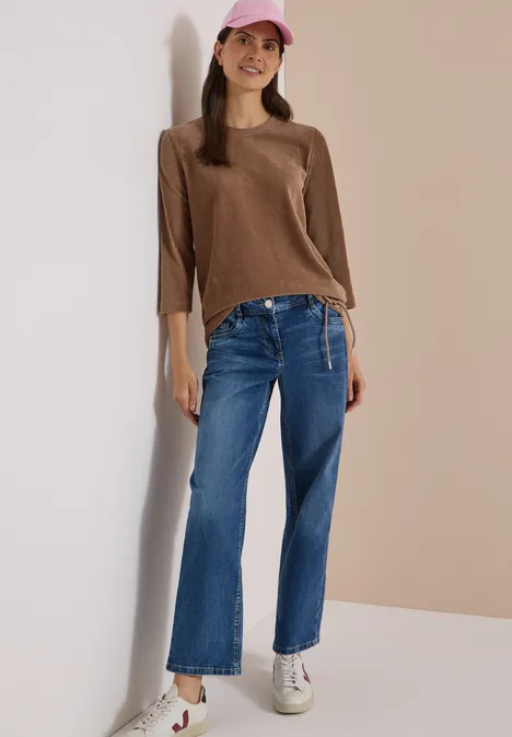 Eine junge Frau lehnt fröhlich an der Wand, trägt braunen Pullover, weite Jeans, weiße Sneaker und rosa Cap.
