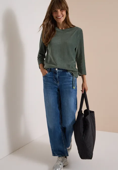 Lächelnde Frau lehnt an Wand, trägt grünes Shirt, weite Jeans und schwarzen Shopper.