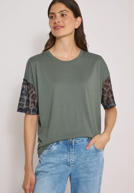 Vrouw in olijfgroen shirt met luipaardprintmouwen en spijkerbroek, hand nonchalant in de zak.