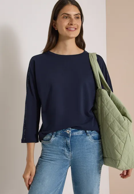 Batwing Shirt mit Leo-Pipings urban dark blue Lächelnde Frau in dunkelblauem Pullover und Jeans mit grüner, gesteppter Umhängetasche vor neutralem Hintergrund.