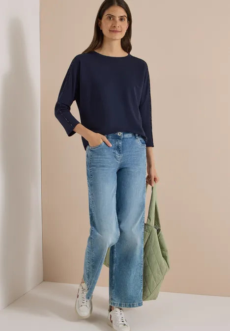 Batwing Shirt mit Leo-Pipings urban dark blue Junge Frau mit dunkelblauem Langarmshirt, hellen Schlagjeans, weißen Sneakern und grüner Tasche vor beigem Hintergrund.