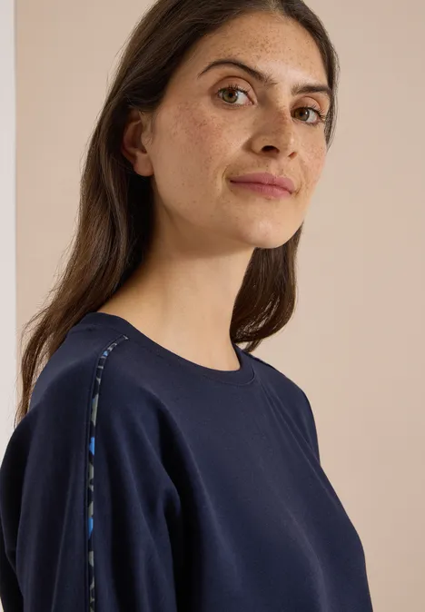 Batwing Shirt mit Leo-Pipings urban dark blue Eine Frau in dunklem Sweatshirt mit dezenten Schulterdetails blickt ruhig zur Kamera.