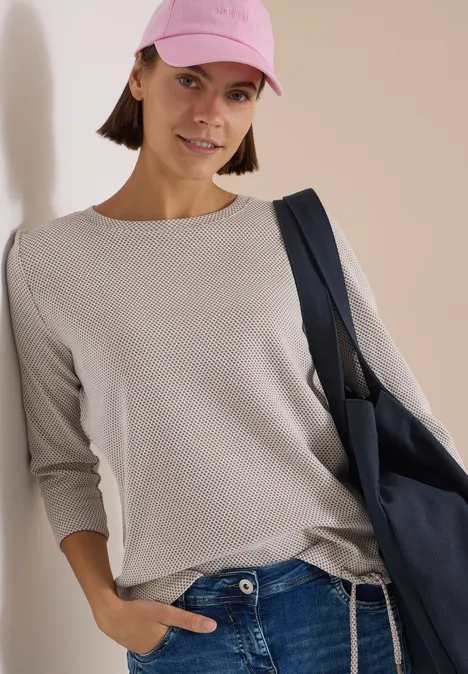 3/4-Arm Shirt mit Rundhals und Struktur natural taupe Frau in beiger Longsleeve, Jeans, rosafarbenem Cap und blauer Tasche lehnt an Wand.