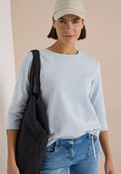 3/4-Arm Shirt mit Rundhals und Struktur easy blue Frau mit Basecap, hellblauem Pullover, Jeans und schwarzer Schultertasche.