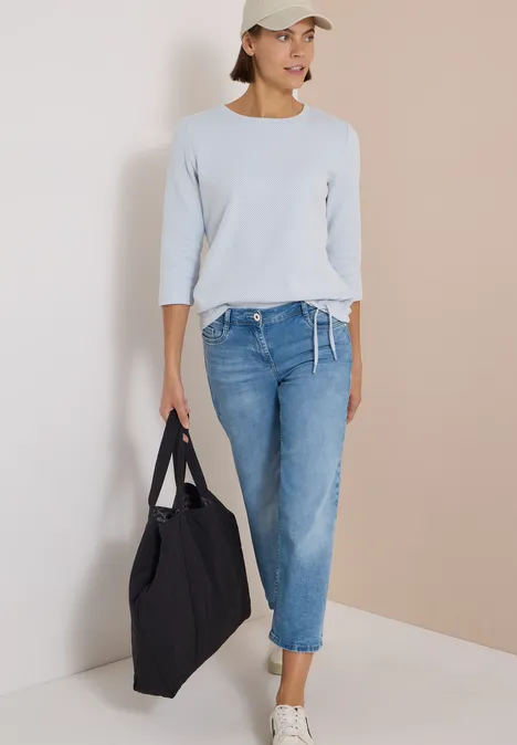 3/4-Arm Shirt mit Rundhals und Struktur easy blue Frau in hellblauem Pullover, Jeans, weißer Cap und großer schwarzer Tasche vor neutralem Hintergrund.