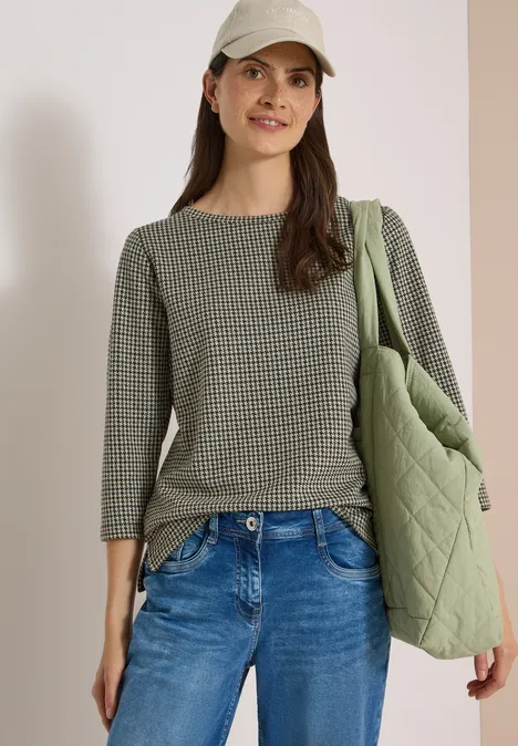 Frau mit graugrünem Pullover, Jeans, Cap und gestepptem Shopper lächelt entspannt