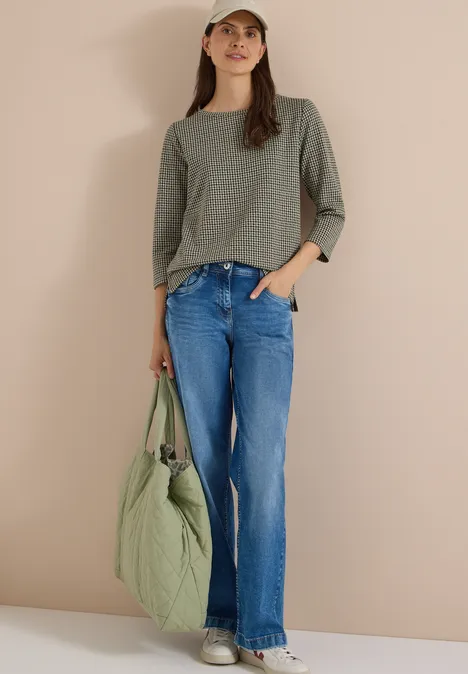 Frau trägt grünes Longsleeve, Schlagjeans und hält eine gesteppte grüne Tasche.