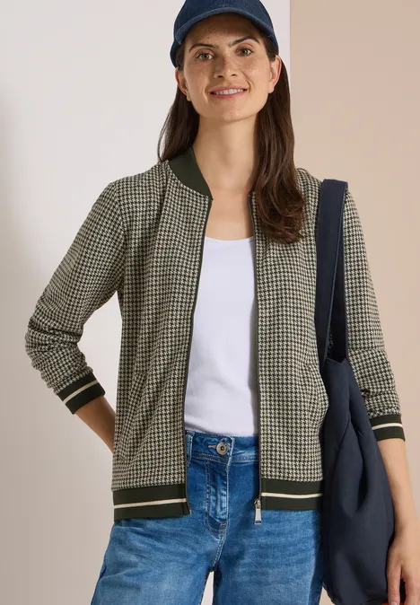 Blouson léger au motif « pepita » raw khaki Frau in kariertem Blouson, weißem T-Shirt, Jeans und Cap mit blauem Shopper lächelnd.