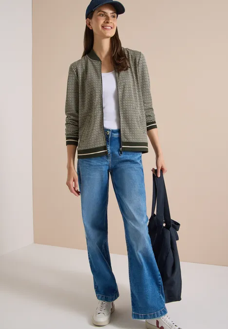 Blouson léger au motif « pepita » raw khaki Eine junge Frau in Jeans, weißem Shirt, khakifarbener Jacke, Sneakers und Cap trägt eine große blaue Tasche.
