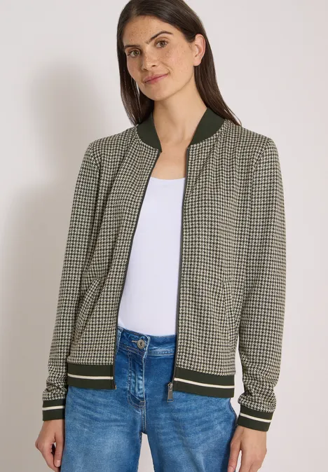 Blouson léger au motif « pepita » raw khaki Frau trägt gemusterte Bomberjacke mit grünen Bündchen, weißes Top und blaue Jeans vor neutralem Hintergrund