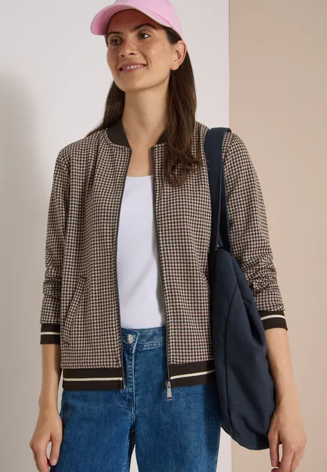Blouson léger au motif « pepita » raw chocolate brown Eine Frau trägt eine karierte Bomberjacke, weißes T-Shirt, Jeans, rosa Cap und eine Schultertasche.