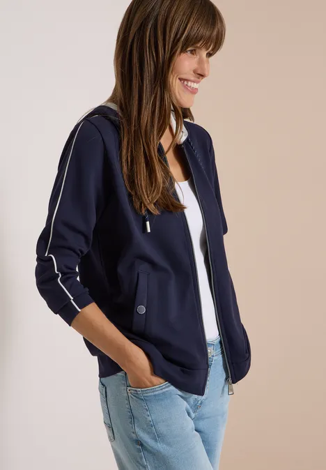 Eine junge Frau trägt eine dunkelblaue Jacke mit Reißverschluss und Jeans, lächelnd zur Seite blickend.