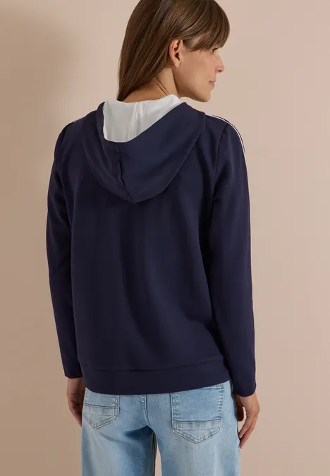 Frau von hinten trägt dunkelblaue Kapuzenjacke mit weißem Innenfutter und Jeans vor beigem Hintergrund.