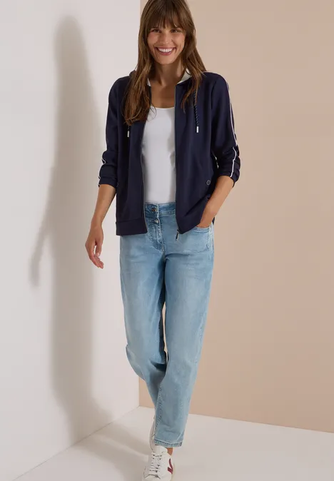 Eine Frau in lässiger Jeans, weißem T-Shirt und marineblauer Jacke, die lächelnd steht.