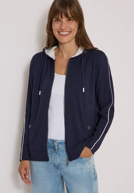 Frau in dunkelblauer Kapuzenjacke, weißem Top und Jeans, lächelnd.