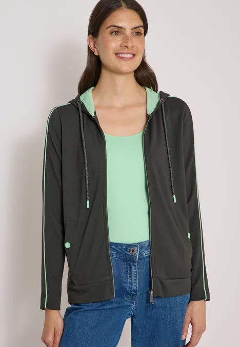 Frau in schwarzer Kapuzenjacke und mintgrünem Top, lächelnd und blickt geradeaus.