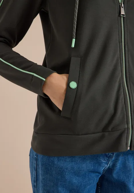 Schwarzer Hoodie mit grünen Paspeln und Knöpfen, Reißverschlussdetails, blaue Jeans, Hand in Tasche