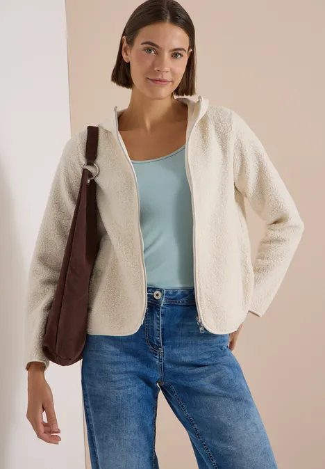 Frau trägt cremefarbene Teddyfleece-Jacke mit Kapuze über türkisfarbenem Top und blauen Jeans, braune Schultertasche.