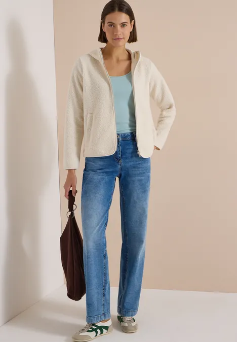 Frau in cremefarbener Jacke, türkisem Top, blauen Jeans und Sneakern hält braune Tasche.