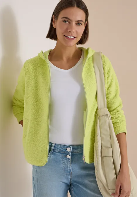 Junge Frau in grüner Fleece-Jacke, weißem Top und Jeans, lächelnd mit Tasche über der Schulter.